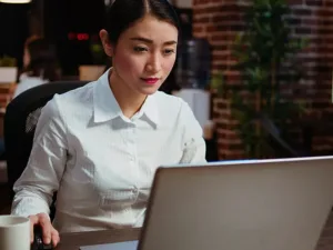 A woman in a white shirt sits at a desk, looking at a laptop screen in an office with brick walls and plants, representing an Association Management Company workspace.
