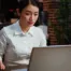 A woman in a white shirt sits at a desk, looking at a laptop screen in an office with brick walls and plants, representing an Association Management Company workspace.